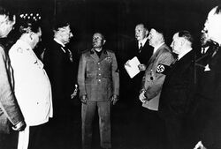 Gruppenbild mit Neville Chamberlain (1869-1940, Premierminister des Vereinigten Königsreichs 1937-1940), Benito Mussolini (1883-1945, Ministerpräsident von Italien 1922-1943) und Adolf Hitler (1889-1945, nationalsozialistischer Diktator Deutschlands 1933-1945) im Fokus. Chamberlains und Hitlers Körpersprache spiegeln sich, während Chamberlains Worte durch den Chefdolmetscher des Auswärtigen Amtes Paul Schmidt (1899-1970) übersetzt werden. (Bayerische Staatsbibliothek, Bildarchiv hoff-20614)