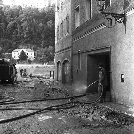 Passau, Michaeligasse: Das Bild zeigt eindrucksvoll die Schlammmassen, die das Donau-Inn-Ilz-Hochwasser in Südostbayern im Juli 1954 in Passau zurückgelassen hat. Der zentimeterdicke Schlamm muss möglichst in noch nassem Zustand entfernt werden, da er nach dem Trocknen mitunter hart wie Beton wird. Nur mit hohem Wasserdruck gelingt es den Helfern, hier der Feuerwehr, den Schlamm zu beseitigen. (Bayerische Staatsbibliothek, Fotoarchiv Fruhstorfer, fruh-08741)