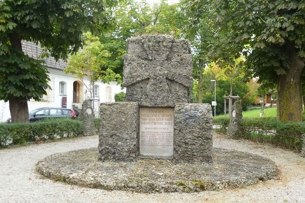 Kriegerdenkmal in Irsee. Das 1936 eingeweihte Denkmal aus einem zentralen steinernen Block mit einem Relief des Eisernen Kreuzes, an dessen Sockel Tafeln mit den Gefallenen des Zweiten Weltkriegs nachträglich angebracht wurden. Die Gefallenen des Ersten Weltkrieges werden auf zehn kleineren Stelen mit je drei Namen wiedergegeben, wie sie links und rechts des zentralen Blocks zu sehen sind. (Fotografie von Bayreuth2009, lizenziert durch CC-BY-SA 3.0 via Wikimedia Commons)