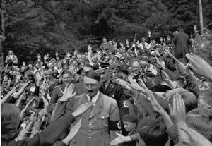 Hitler als "volksnaher Führer" beim Bad in der Menge. Anhand einzelner Details (wie dem Fotografen, der im Hintergrund auf einer Leiter steht) und unveröffentlichten Aufnahmen der zugehörigen Bildserie erkennt man, dass solche Aufnahmen sorgfältig inszeniert wurden. Fotografie von Heinrich Hoffmann, August 1933. (Bayerische Staatsbibliothek, Bildarchiv hoff-8153)