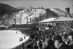 Das Olympia-Skistadion mit dem Olympia-Haus, Fahnen der teilnehmenden Nationen und voll besetzter Tribüne. Fotografie von August Beckert von 1936. (Bayerische Staatsbibliothek, Bildarchiv ansi-005724)